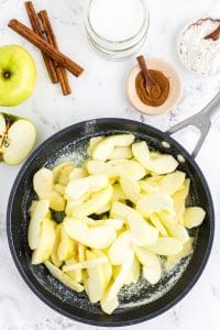 Frying pan with apple slices, bowl with ground cinnamon, cinnamon sticks, bowl with flour, an apple on marble countertop
