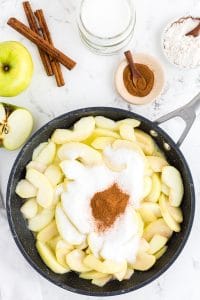 Frying pan with apple slices, sugar and ground cinnamon, bowl with ground cinnamon, cinnamon sticks, bowl with flour, an apple on marble countertop