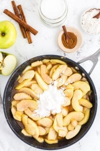 Frying pan with apple mixture and flour, bowl with ground cinnamon, cinnamon sticks, bowl with flour, an apple on marble countertop