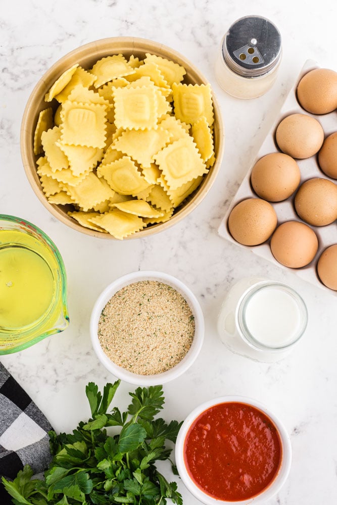 Individual ingredients for fried raviolis:Bowl with uncooked raviolis, jar with parmesan, tray of eggs, jar filled with buttermilk, bowl with bread crumbs, bowl with Marinara Sauce, glass pitcher, black and white checked linen, parsley.