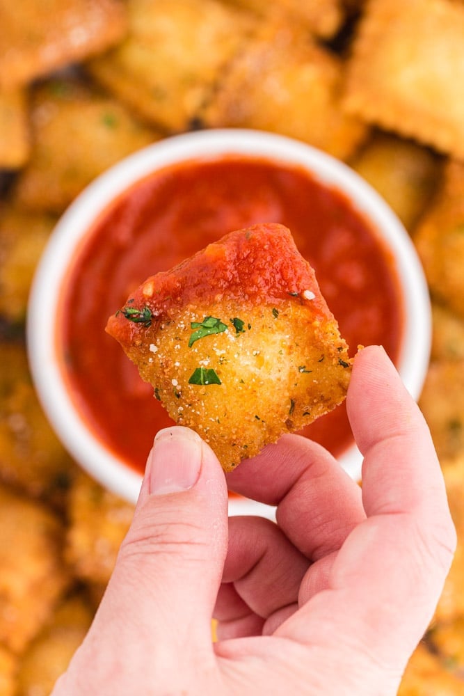Hand holding a fried ravioli partially covered with Marinara Sauce, above a baking tray covered with fried raviloi.