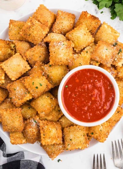 Plate filled with Fried Ravioli and a white bowl with Marinara Sauce, black and white checked linen ,parsley, 3 forks on a marble countertop