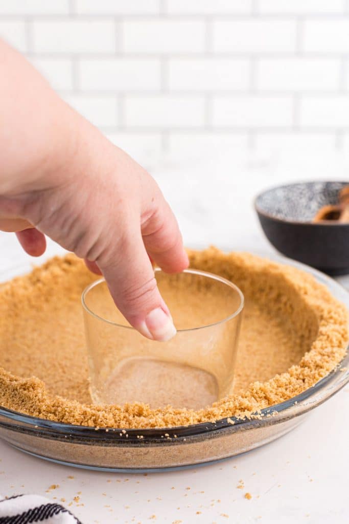 water glass pressed on ingredients for Graham Cracker Pie Crust in pie dish, bowl with sugar, white subway tiles in the back