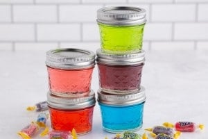 Five glasses with lid filled with different colored Jolly Rancher Candy dissolved in vodka, jolly rancher candy, white subway tiles in the background