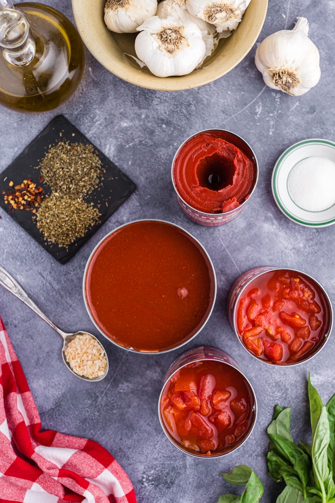 individual ingredients for marinara sauce: canned tomatoes, tomato sauce, tomato paste, herbs, olive oil, garlic, sugar, and fresh herbs