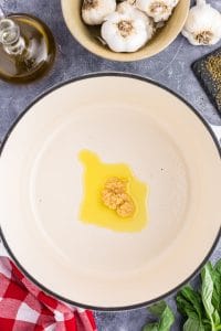 White Dutch Oven with handles filled chopped garlic sautéing in vegetable oil, red and white checked linen, basil, garlic, glass bottle with vegetable oil