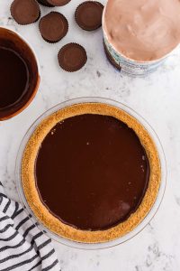 Glass pie dish with graham cracker crust filled with chocolate ganache, peanut butter cups, peanut butter chocolate ice cream, bowl with chocolate ganache, grey and white striped linen on marble counter top