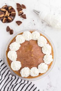 Glass pie dish with graham cracker crust filled with peanut butter chocolate ice cream decorated with whipped cream , bowl with cut peanut butter cups, grey and white striped linen on marble counter top