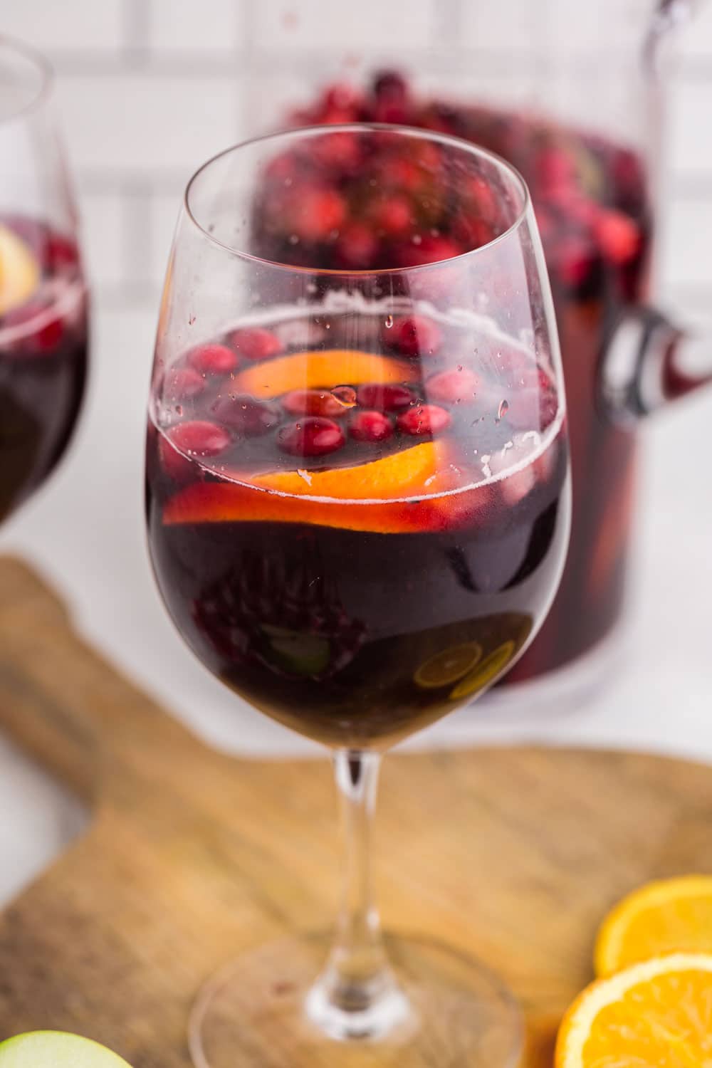 Two wine glasses filled with Red Sangria on a wooden kitchen board, sliced orange, glass pitcher in the background, with white subway tiles.