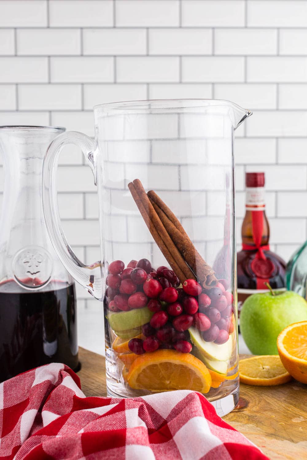 Glass pitcher filled with cranberries, cinnamon sticks, sliced apples and oranges, a glass decanter with red wine and half an orange on a wooden kitchen board, red and white checked linen, a bottle of Grand Marnier, white subway tiles as background.