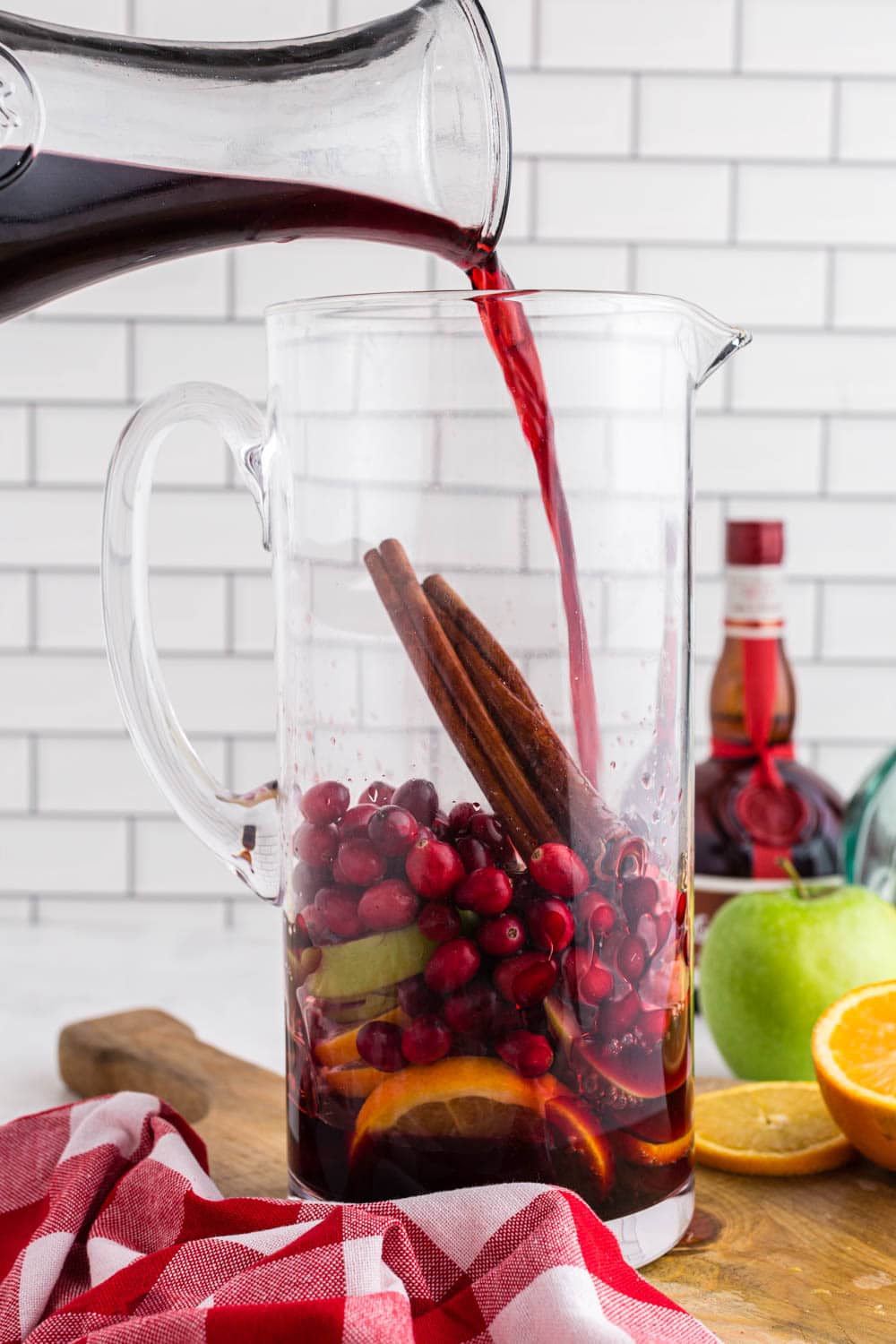 Glass decanter filled with red wine getting emptied into a glass pitcher filled with cranberries, cinnamon sticks, sliced apples and oranges, half an orange on a wooden kitchen board, red and white checked linen, a bottle of Grand Marnier, white subway tiles as background.