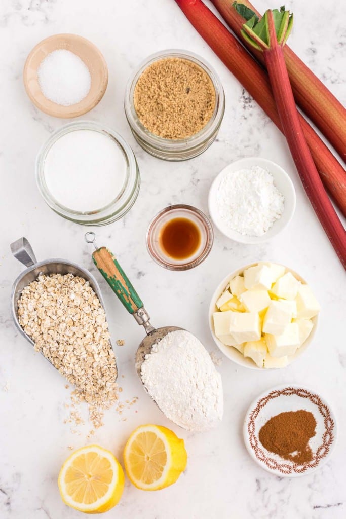 Several bowls, spoons and plates with Rhubarb Crisp ingredients on marble countertop