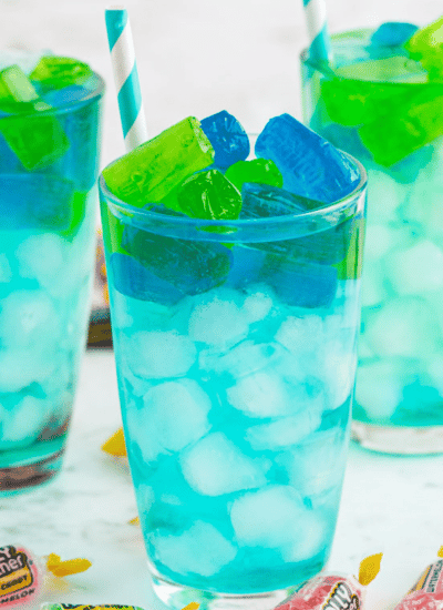 Three glasses of turquoise Jolly Rancher Cocktail with paper straws, jolly rancher candy on marble countertop