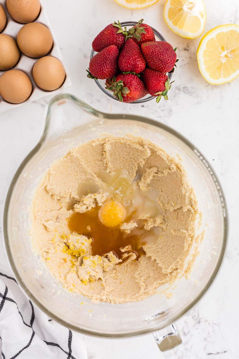 Mixing bowl with cake batter and eggs, strawberries, lemon, eggs on a marble countertop