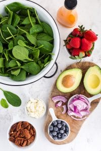 Bowl filled with baby spinach, bowl with pecans, avocado cut in half, bowl of blueberries, bowl of cut onions, bowl of feta cheese, bowl with strawberries, half , a wooden kitchen board, bottle with salad dressing on a marble countertop
