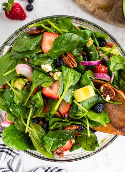 Strawberry Spinach Salad with wooden serving spoon, bowl of strawberries, half an avocado , glass with salad dressing, black and white checked linen on marble countertop