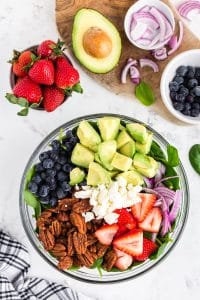 Glass Bowl filled with spinach, pecans, cut strawberries, cut avocado, blueberries, onions, feta cheese, bowl with strawberries, half an avocado, bowl with blueberries, bowl with onions, black and white checked linen on a white marble countertop