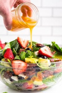 Salad dressing getting poured into a glass bowl filled with Strawberry Spinach Salad, white subway tiles in the back