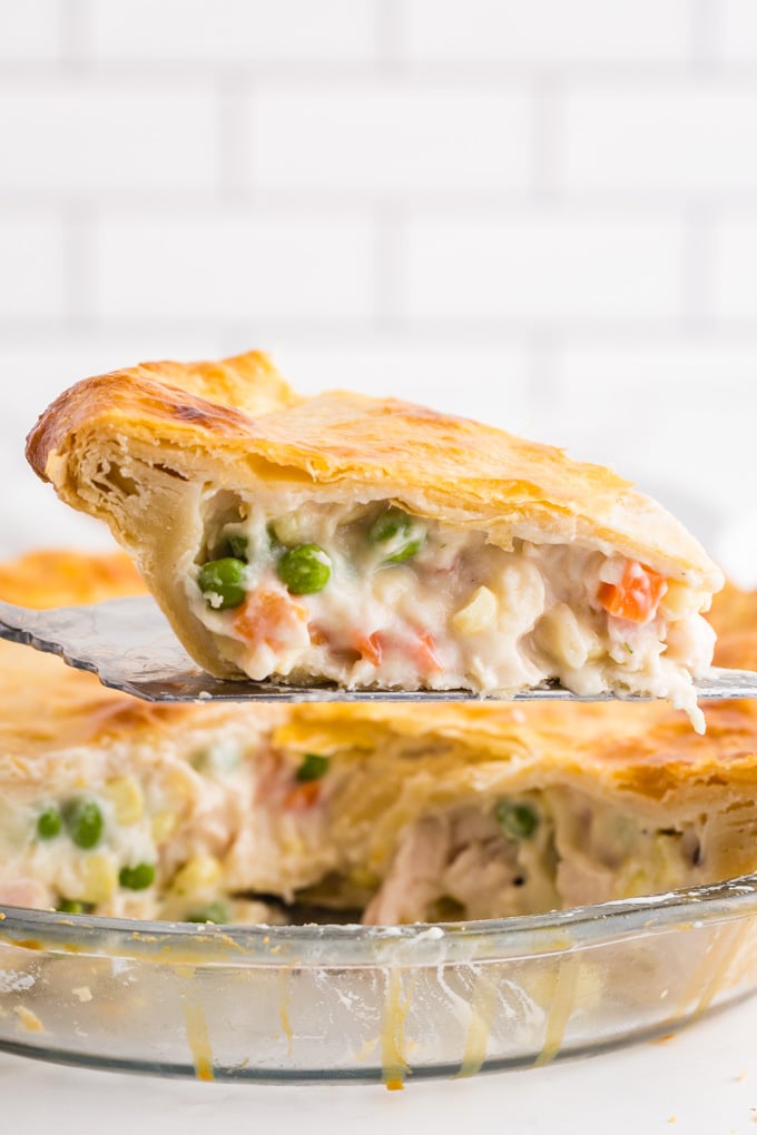 Slice of Chicken Pot Pie on a silver serving spoon, baked chicken pot pie in pie dish on a marble countertop, white staked tiles in the background.