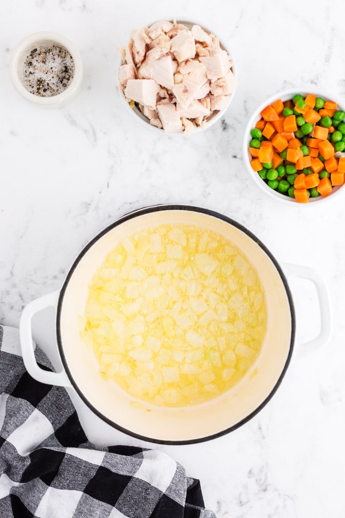 Dutch oven with melted butter and sauteed onions, bowl with frozen peas and carrots, bowl with chicken, bowl with spices, black and white checked linen on marble countertop