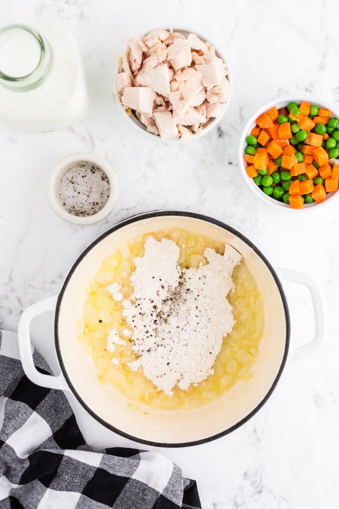 Dutch oven with melted butter, sauteed onions, flour and spices, bowl with frozen peas and carrots, bowl with chicken, bowl with spices, black and white checked linen on marble countertop.