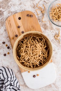Bowl with melted chocolate mix and chow mein noodles on a wooden kitchen board, bowl with chow mein noodles, black and white striped linen on a marble countertop