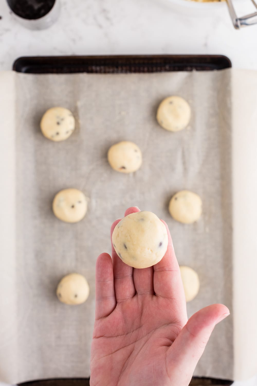 Rolled cookie dough ball held over a baking tray lined with parchment paper filled with rolled balls of cookie dough, metal measuring spoon filled with chocolate chips, striped kitchen linen