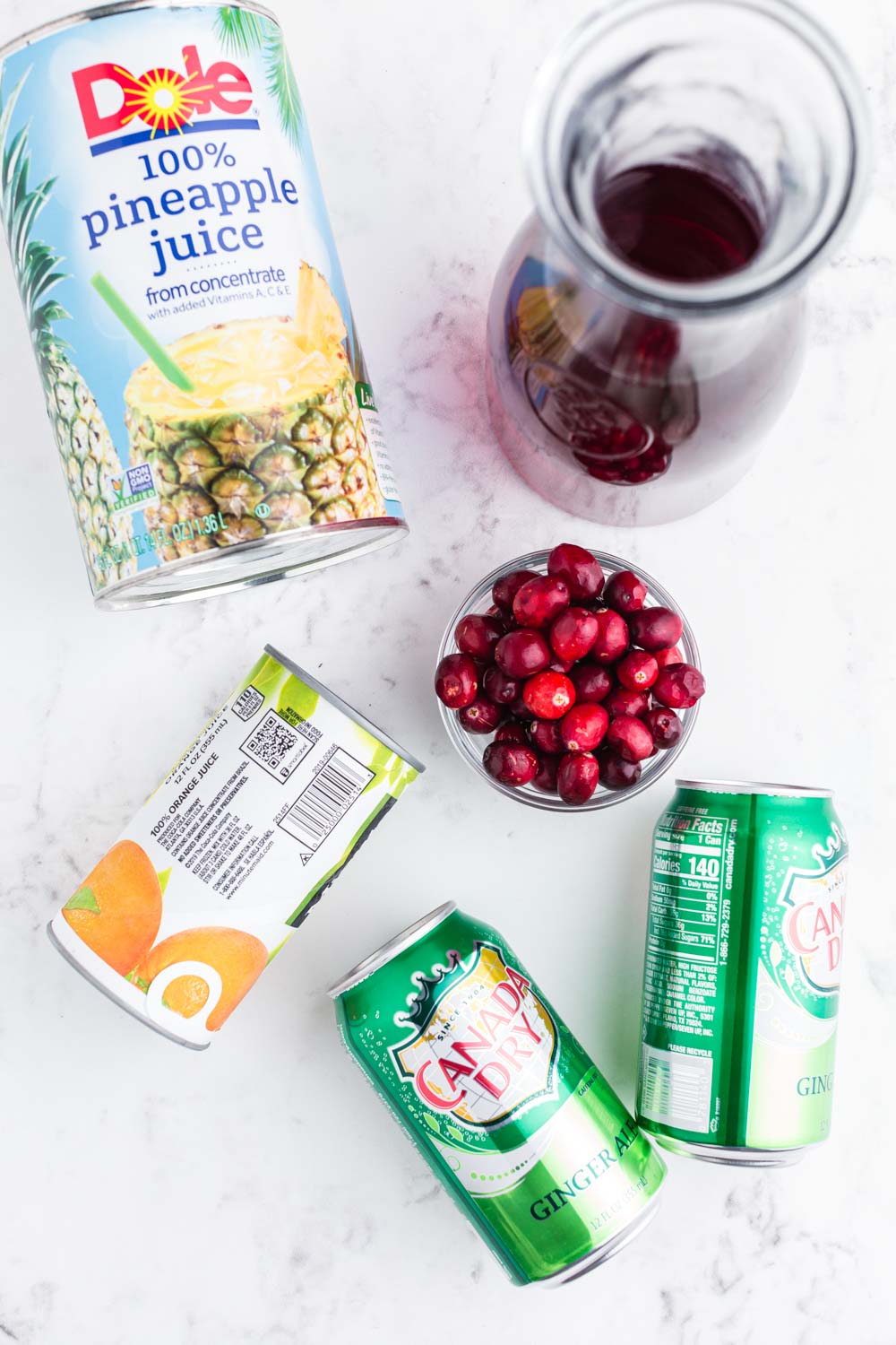 Can of frozen orange juice concentrate, 2 cans of ginger ale, bowl with cranberries, pitcher with cranberry juice, can of pineapple juice, on marble countertop.