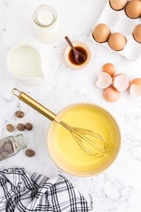 Egg yolk sugar in mixture with whisk in mixing bowl, pitcher with whipping cream, cracked egg shells, bowl with vanilla extract, nutmeg, black and white striped linen on marble countertop