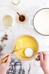 Mixture of whipping cream with nutmeg scooped into egg yolk mixture, pitcher with whipping cream, bowl with vanilla extract, nutmeg, black and white striped linen on marble countertop