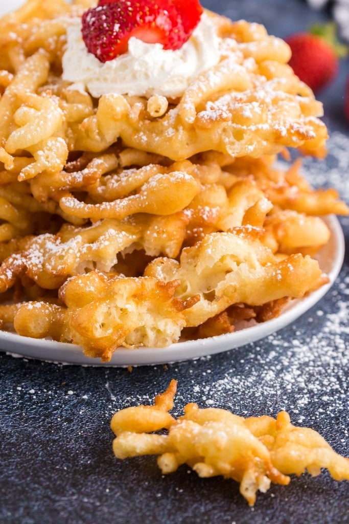 funnel cakes stacked on a plate with powdered sugar and whipped cream and strawberries on toop