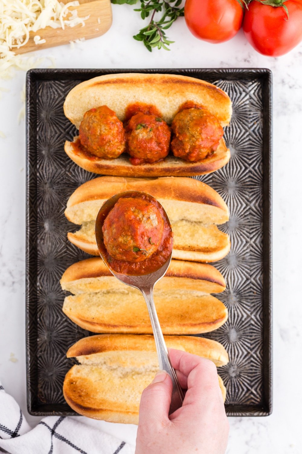 Spoon with meatball held over cookie sheet with sandwich bun, one sandwich bun filled with three meatballs, tomatoes, basil, shredded cheese, on a marble countertop.