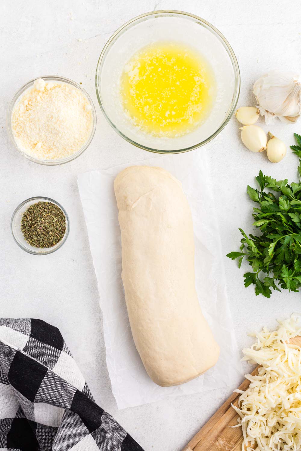 Bread dough on parchment paper, grated mozzarella on wooden kitchen board, fresh parsley, whole garlic cloves, bowl of melted butter, bowl of grated parmesan cheese, small bowl of Italian seasoning, black and white checkered linen, on a white marble surface. 
