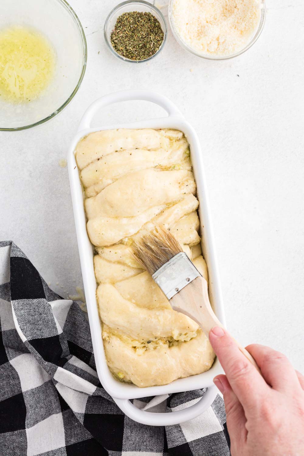 Unbaked pull apart garlic bread in a white loaf baking dish being brushed with melted butter, bowl of melted butter, bowl of seasoning, bowl of grated cheese, black and white checkered linen, on a white marble countertop.