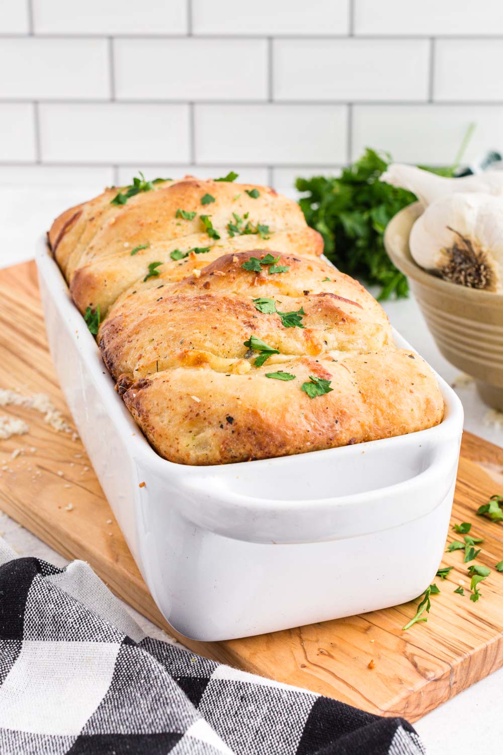 Pull Apart Garlic Bread in a bread loaf baking dish on wooden kitchen board, bowl of whole garlic, fresh parsley, black and white checkered linen, white stacked tiles in the background. 