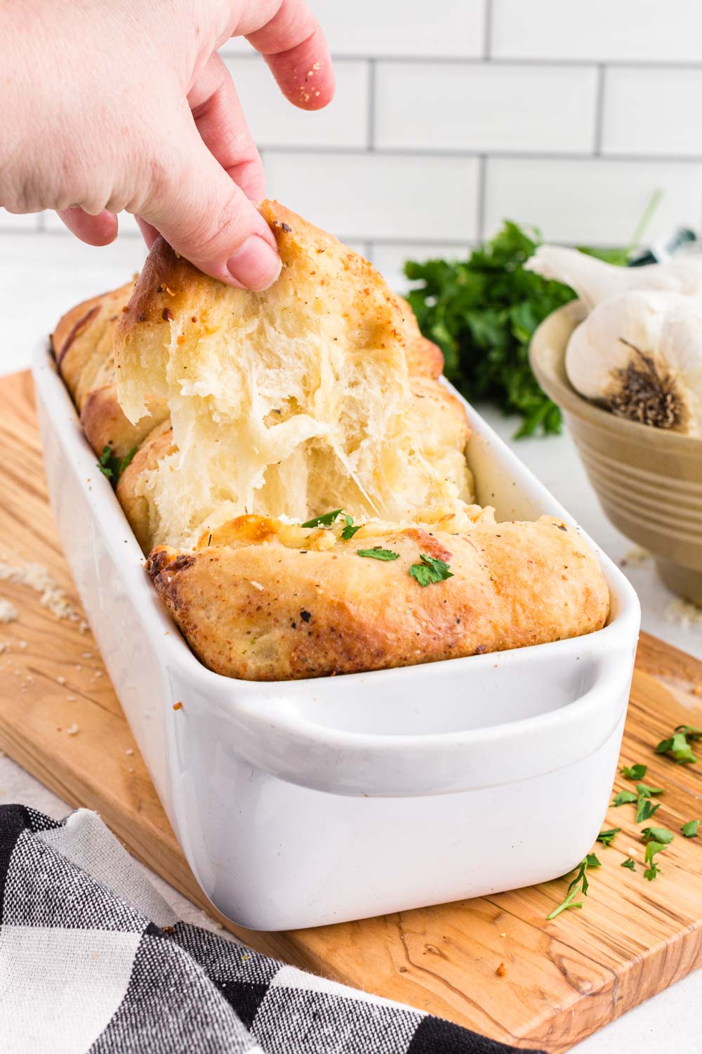 Hand pulling on Pull Apart Garlic Bread in white bread loaf baking dish on wooden kitchen board, bowl of garlic cloves, fresh parsley, black and white checkered linen, on a white marble surface, white stacked tiles in the background.