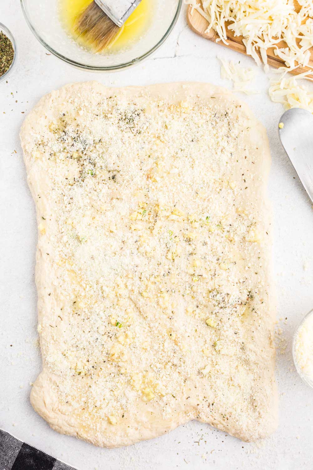 Rolled out bread dough topped with seasoning, minced garlic, melted butter, and grated cheese, on a white marble surface.