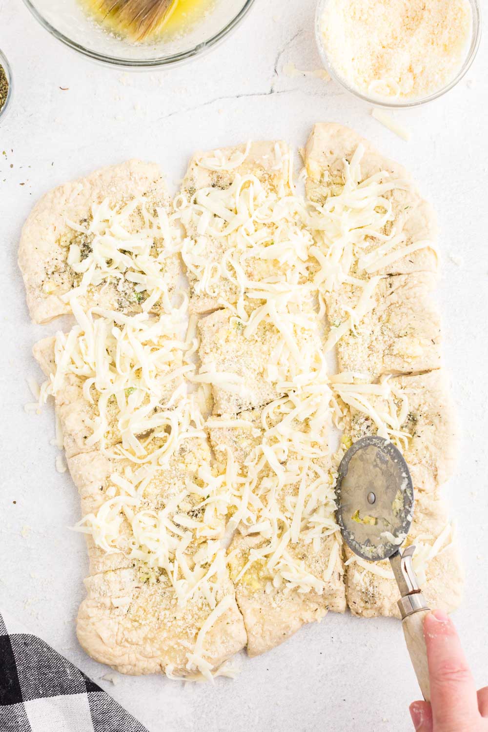 Bread dough with seasoning and grated cheese, pizza cutter slicing squares, on a white marble surface.