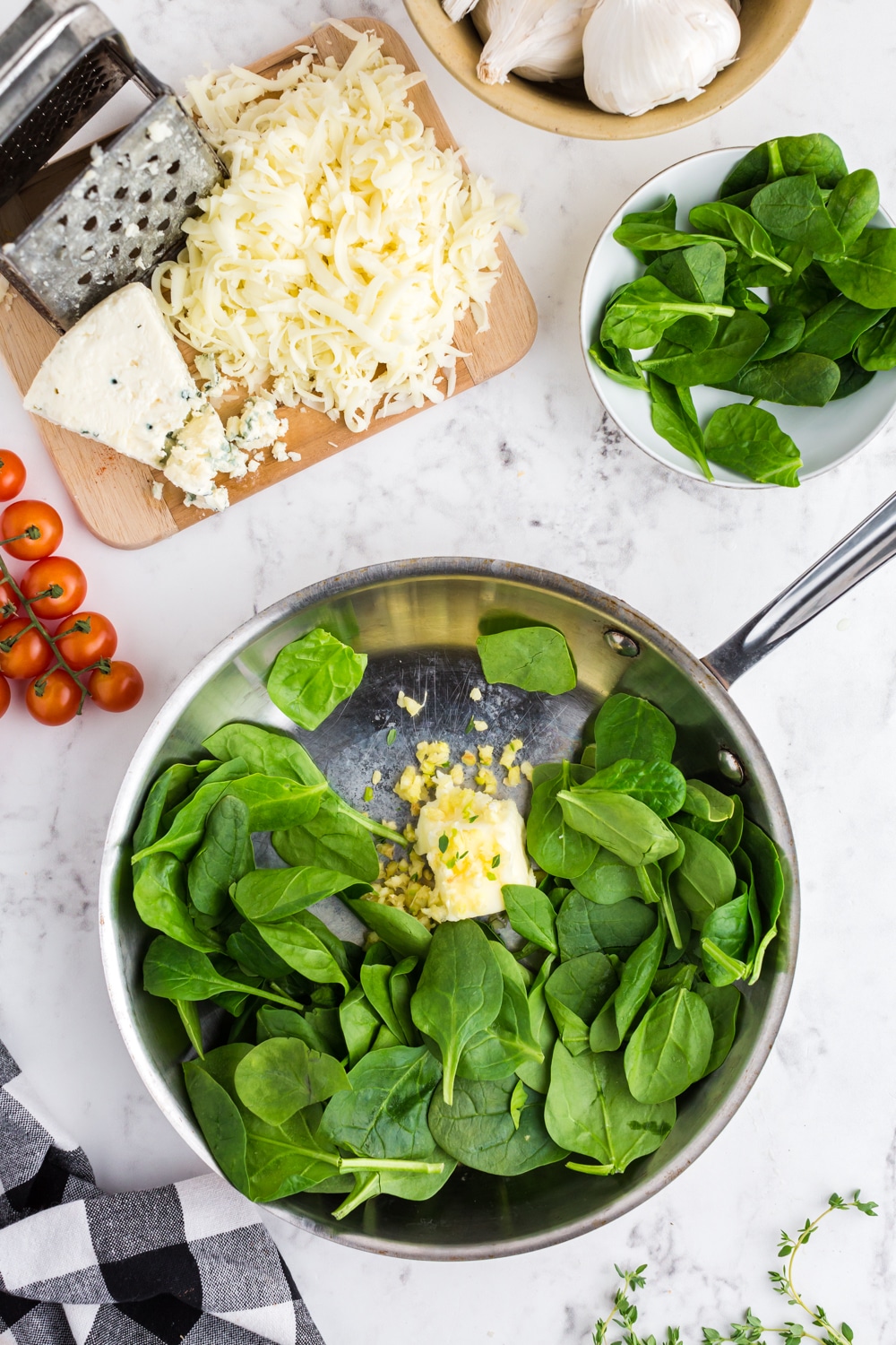 Spinach Garlic and Butter in a Skillet, cheese, grated cheese, grater on a wooden kitchen board, bowl with spinach leaves, cherry tomatoes, black and white checked kitchen towel, bowl of garlic cloves, on marble countertop