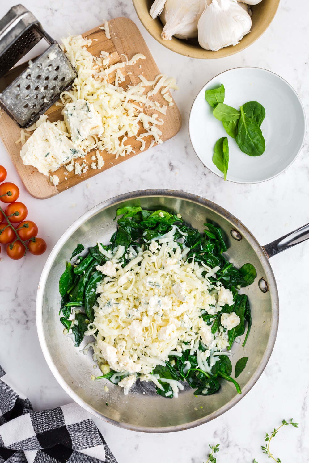 Spinach Garlic Butter and grated cheese in a Skillet, cheese, grated cheese, grater on a wooden kitchen board, bowl with spinach leaves, cherry tomatoes, black and white checked kitchen towel, bowl of garlic cloves, on white marble countertop