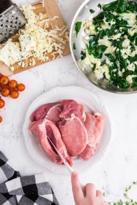 Knife cutting a slit in a pork chop, plate with pork chops, spinach, garlic, butter and grated cheese in a Skillet, cheese, grated cheese, grater on a wooden kitchen board, cherry tomatoes, black and white checked kitchen towel on white marble countertop