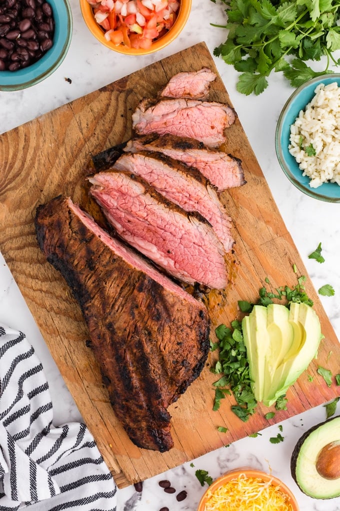Cooked and sliced Chipotle Steak on wooden kitchen board, sliced avocado parsley, salad ingredients in separate bowls, black and white striped linen cloth, on marble countertop