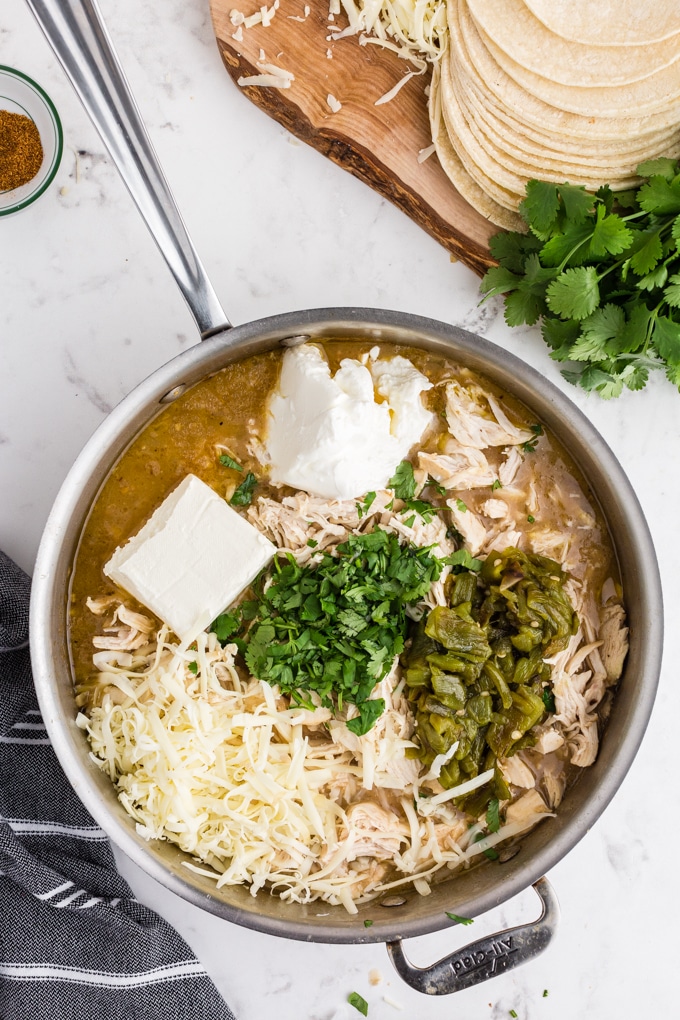 Skillet with sauce, chicken breast, jalapenos, parsley, cream cheese, striped linen, fresh cilantro and grated cheese on a wooden board, on marble countertop