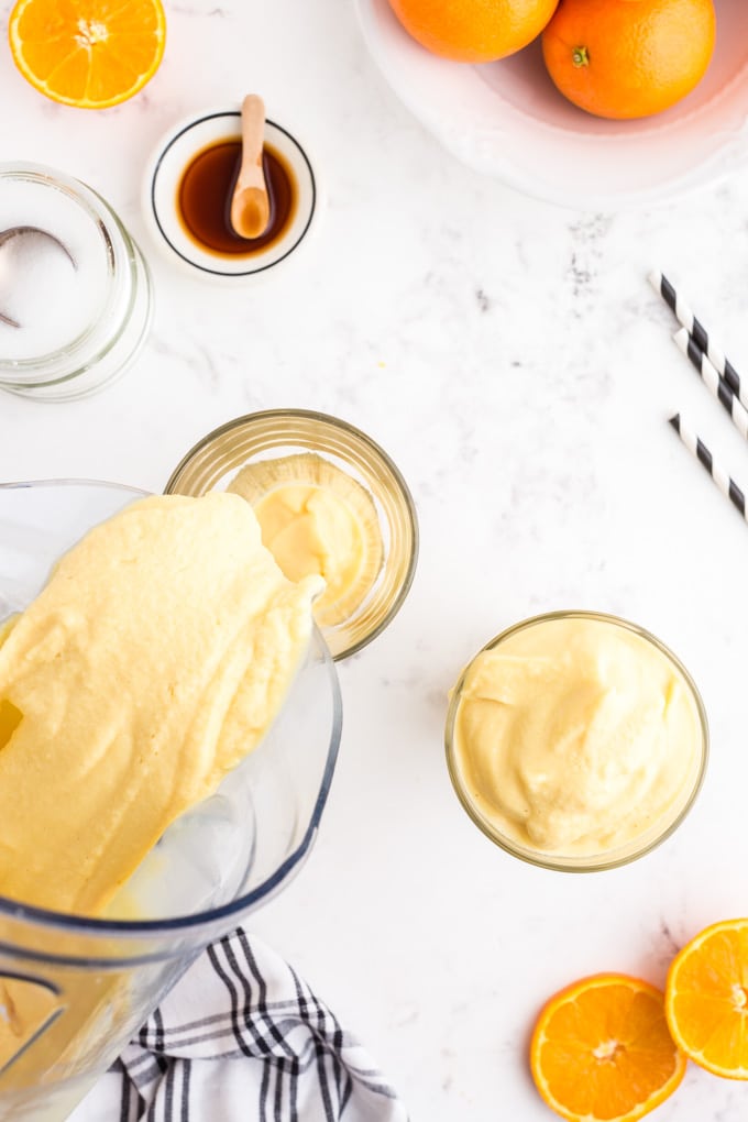 Pouring mixed Orange Julius ingredients into two glasses, remaining ingredients on the countertop.