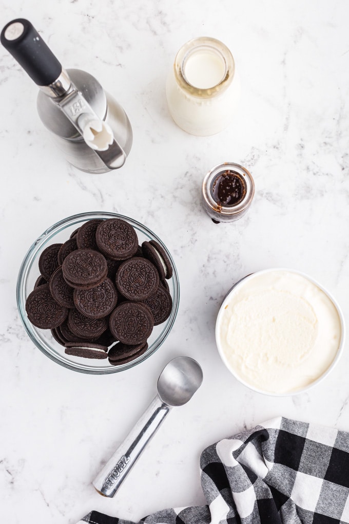 Bowl of Oreo cookies, tub of vanilla Ice cream, metal Ice cream scoop, bottle of chocolate sauce syrup, cream, black and white checked linen, on marble countertop