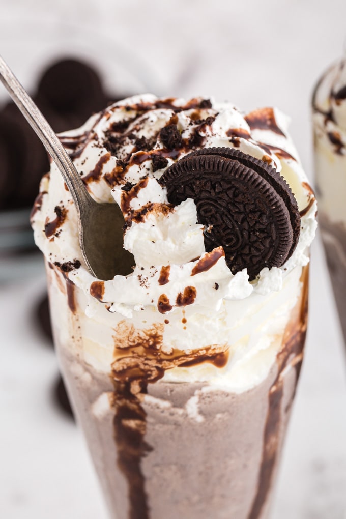 Oreo milkshake in a glass decorated with whipped cream, an Oreo cookie, and chocolate syrup drizzle, metal spoon in the milkshake, bowl of Oreo cookies in the background, on a marble countertop