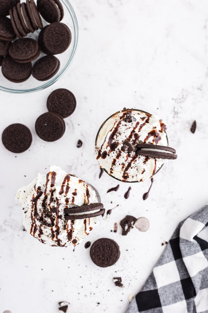 Above shot of two glasses of Oreo milkshake decorated with whipped cream, chocolate drizzle, and Oreo cookies, glass bowl of Oreo cookies, black and white checked linen, on marble countertop