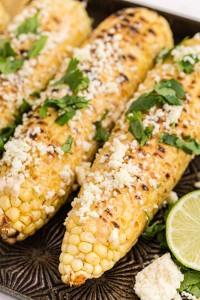 close up of Mexican Street Corn with garnish, on baking sheet, lemons on side