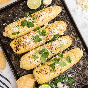 Mexican Street Corn with garnish, on baking sheet, lemons on side, striped linen cloth, atop a white marble surface