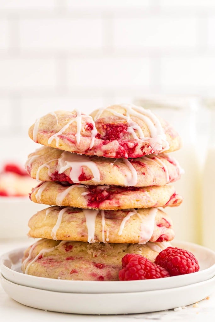 lemon raspberry cookies stacked on a white dish with fresh raspberries as garnish and glasses of milk in the background